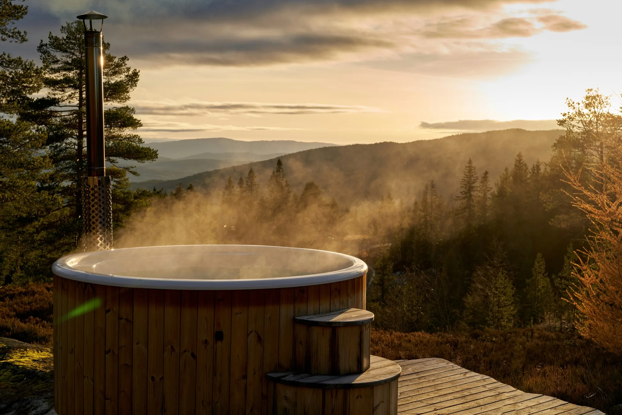 Bain nordique en bois fumant sur une terrasse face à une vallée boisée au coucher du soleil